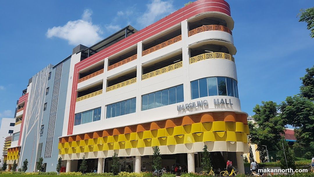 A multi-story building with a colorful facade, featuring a parking garage and retail spaces. The building has a white base with decorative yellow and orange wave-like patterns on the lower levels. The upper levels have balconies with red and yellow railings. The name "MARSLING MALL" is visible in large silver lettering on the white facade. The sky is blue with some clouds.