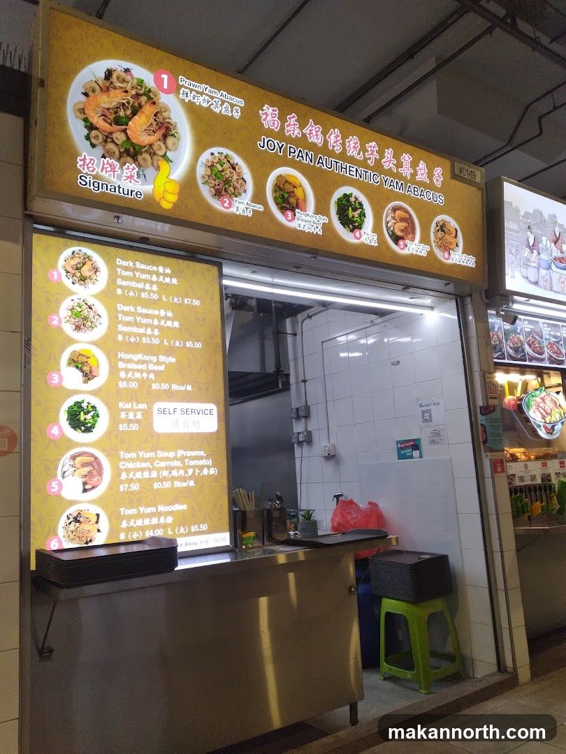 A brightly lit food stall with a large illuminated sign displaying menu items and the business name. Below the main sign, a vertical illuminated menu board lists various dishes with prices. The stall counter is visible, with some kitchen equipment and supplies.