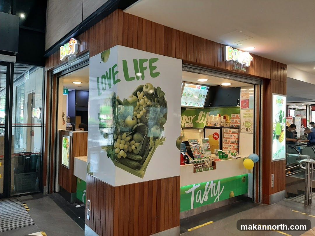 The exterior of a Boost Juice Bar storefront. The shop has a wooden facade with a prominent "Boost" logo above the entrance. A large graphic on the side of the building features the text "LOVE LIFE" and an illustration of fruits and vegetables. The counter area is visible, with menu boards and products displayed.