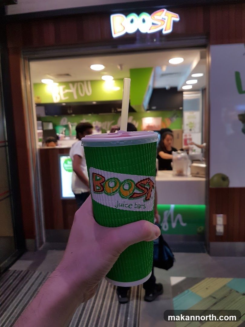 A hand holds a green cup with the Boost Juice Bars logo. In the background, the storefront of a Boost Juice bar is visible, with the "Boost" logo illuminated above the entrance. Inside, staff members are seen behind the counter.