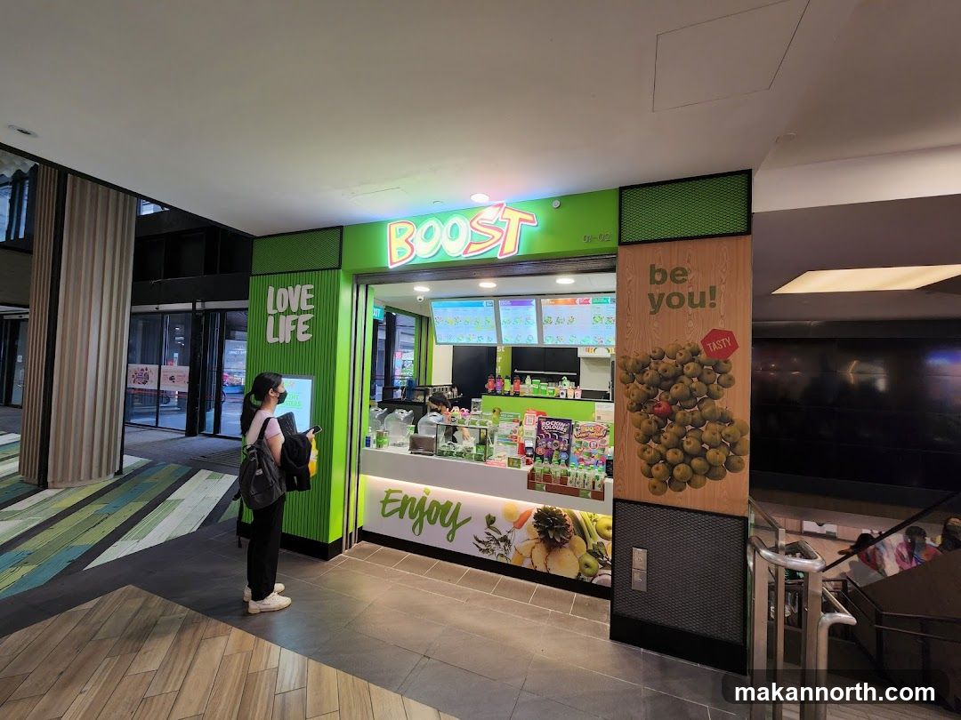 A woman stands in front of a Boost Juice Bar storefront. The store has a bright green facade with the "BOOST" logo prominently displayed. To the left, a sign reads "LOVE LIFE". To the right, a large vertical advertisement features apples and the text "be you!". The interior of the store, including the counter and menu boards, is visible through the open entrance.
