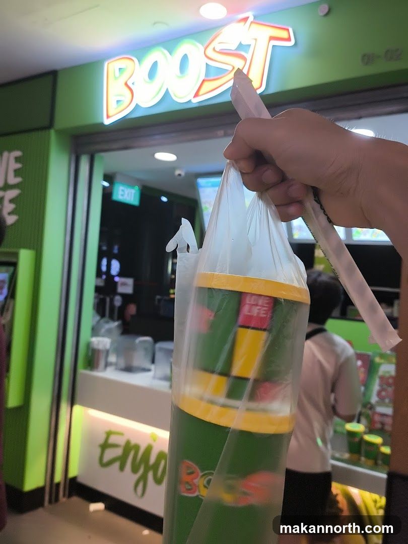 A hand holds a plastic bag with several drinks inside, in front of a Boost Juice Bar storefront. The store has a bright green facade with the "BOOST" logo illuminated in neon letters above the entrance. An "EXIT" sign is visible inside.