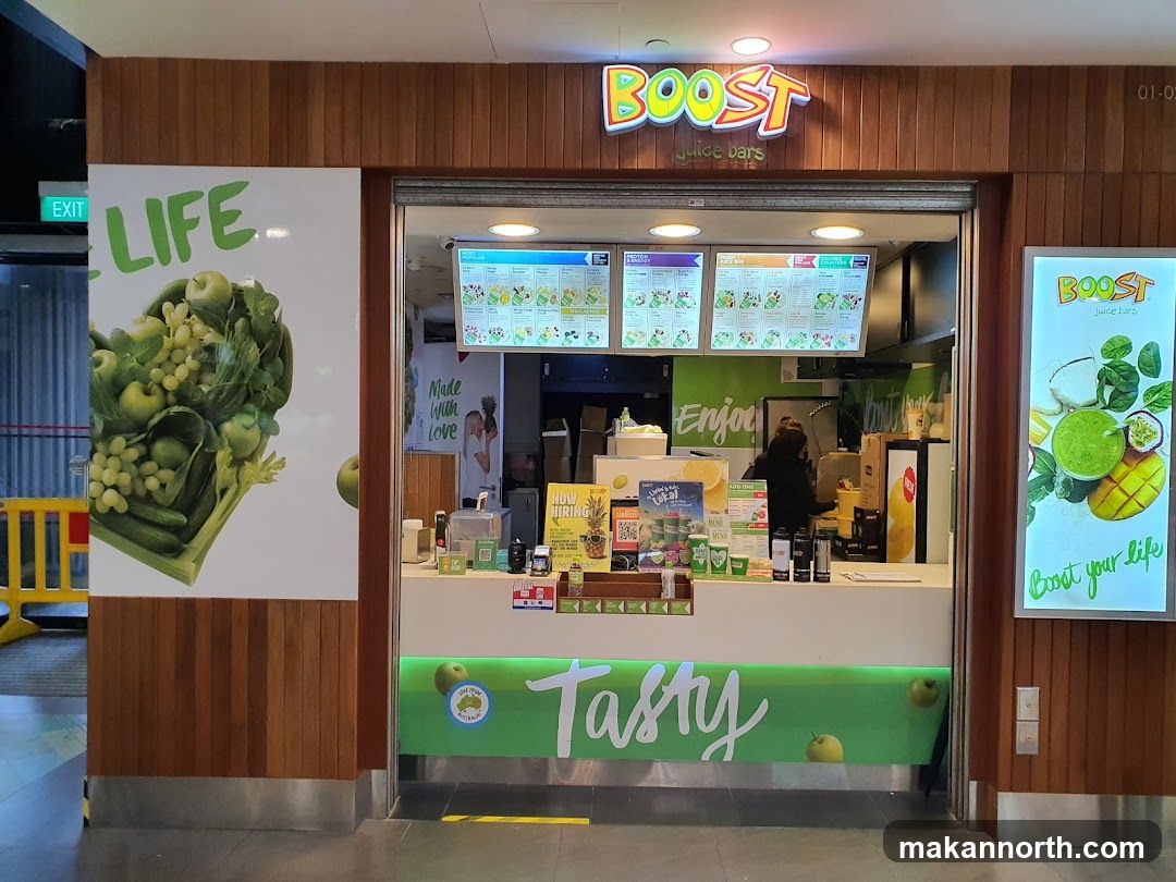 The exterior of a Boost Juice Bars shop with its prominent signage and menu boards visible. The shop features a clean, modern design with wooden paneling and bright green accents. A large graphic of fruits and vegetables is displayed on the left, and a glowing advertisement for Boost Juice Bars is on the right. The entrance is open, revealing the counter and staff inside.