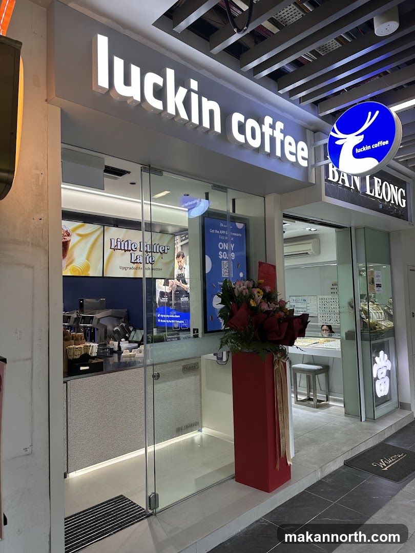 The exterior of a Luckin Coffee shop with its prominent illuminated signage. Through the glass doors, a counter area with coffee machines and displays is visible. A large floral arrangement in a red planter stands outside the entrance.