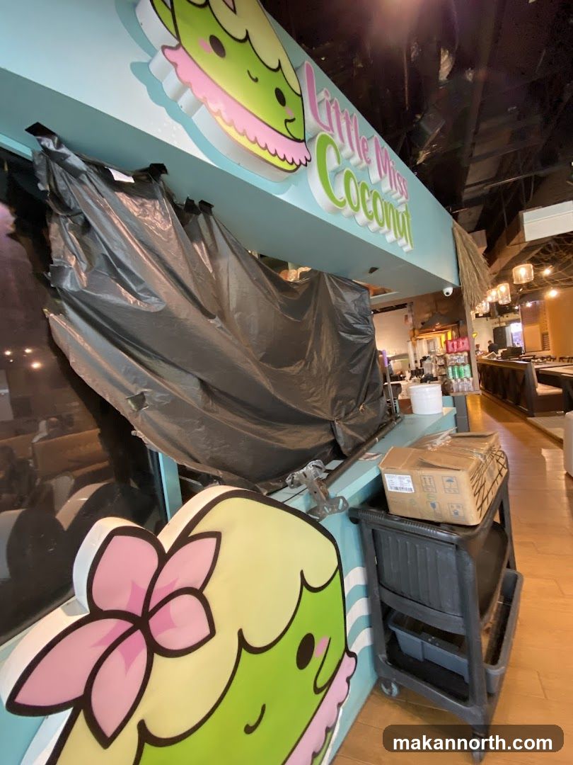 The exterior of a shop with a sign that reads "Little Miss Coconut" and features a cartoon character. The storefront is partially covered with black plastic sheeting. A rolling cart with boxes is visible to the right.