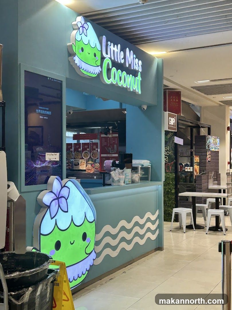 A bright, light blue storefront with the name "Little Miss Coconut" displayed in neon green lettering above the counter. A cartoon coconut character with a flower on its head is featured prominently on the left side of the storefront. A digital menu board is visible on the left, displaying text and images. The counter area has various items displayed, and seating areas with tables and stools are visible in the background.