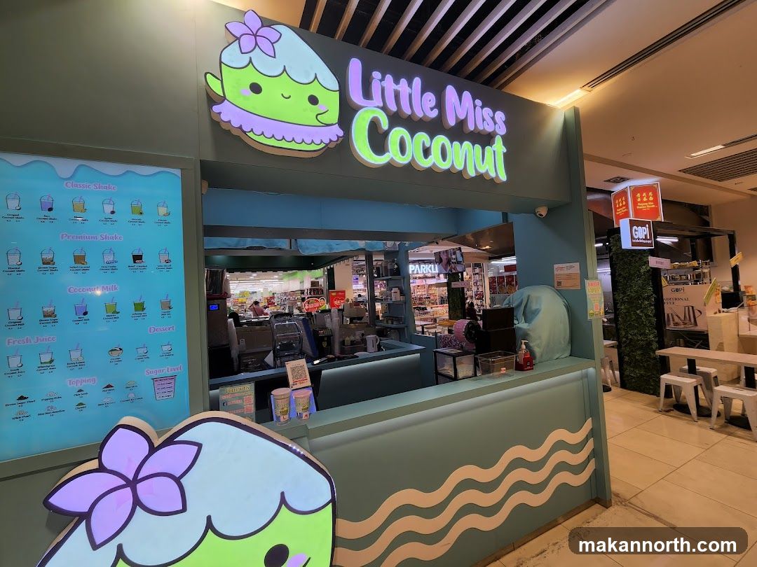 A brightly lit shop front for "Little Miss Coconut" with a large cartoon character logo and the shop name in neon letters. A menu board is visible on the left, displaying various shakes, coconut milk drinks, fresh juices, desserts, and toppings with prices. The counter area is clean and modern, with some equipment visible behind it.