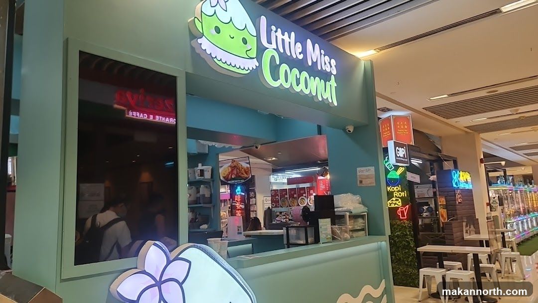 A light green storefront with a prominent sign that reads "Little Miss Coconut" with a cartoon coconut character. The storefront has a counter and display cases. To the left, a dark reflective surface shows a reversed neon sign for "GRANITE CAFE". To the right, there are tables and chairs outside the shop, and further down the mall, arcade games are visible.