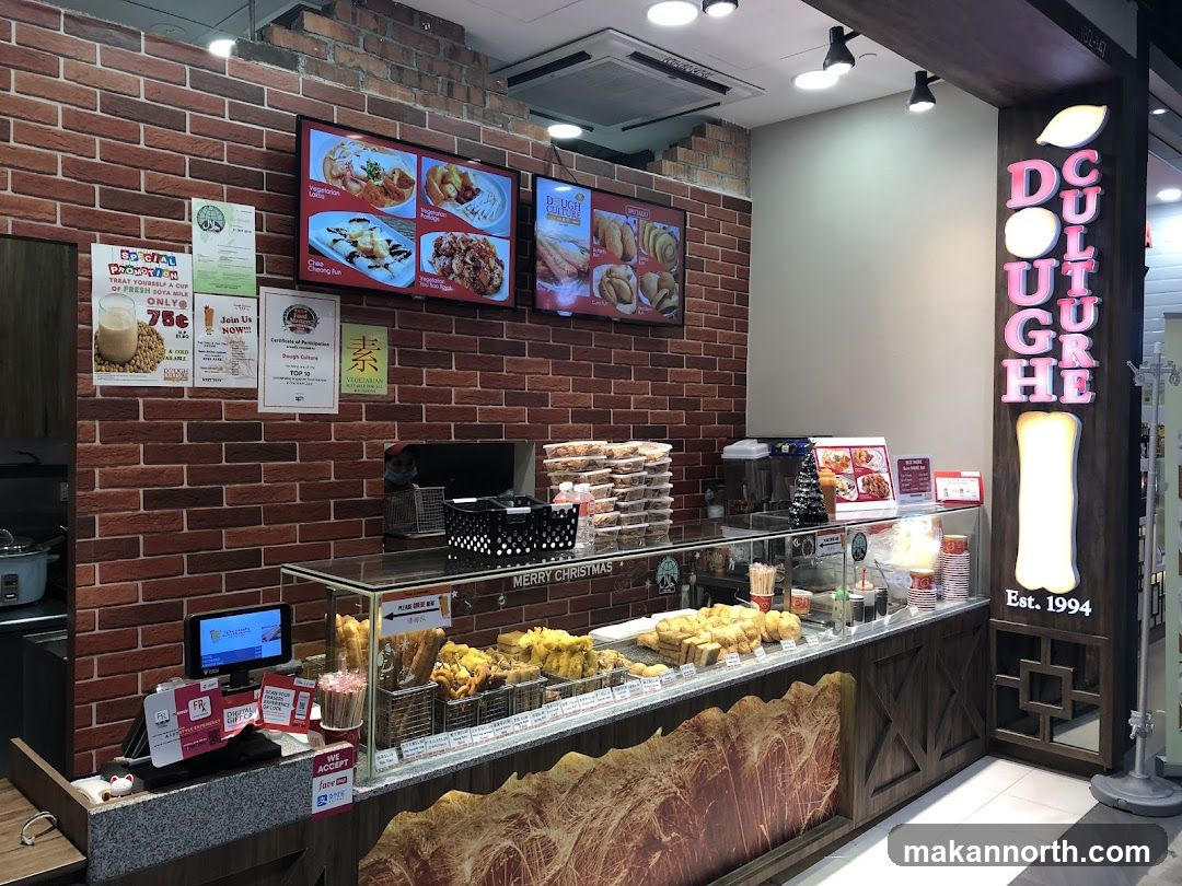 The exterior of a food establishment with a brick wall interior. A display counter showcases various fried food items and pastries. Digital screens display food images and promotions. The business name "DOUGH CULTURE" is prominently displayed on a vertical sign.