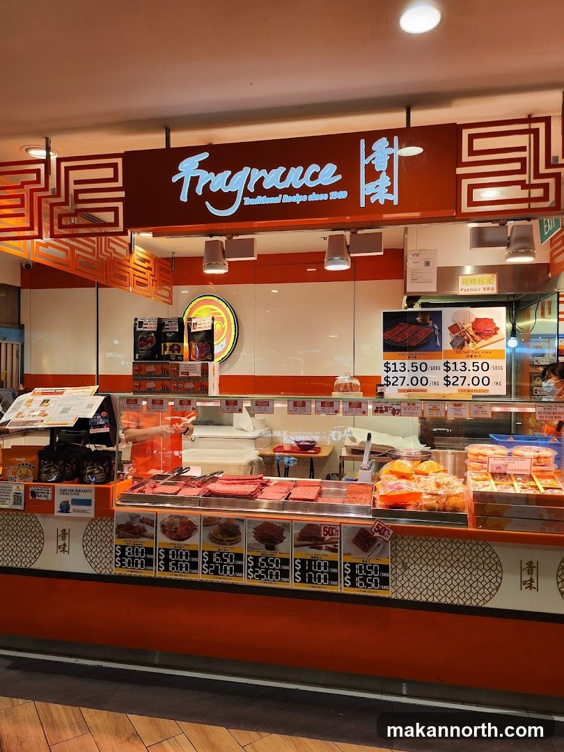 A brightly lit food stall with a prominent orange and white facade. The stall displays various types of dried meat products, with prices listed below. The brand name "Fragrance" is displayed in large, illuminated lettering at the top.