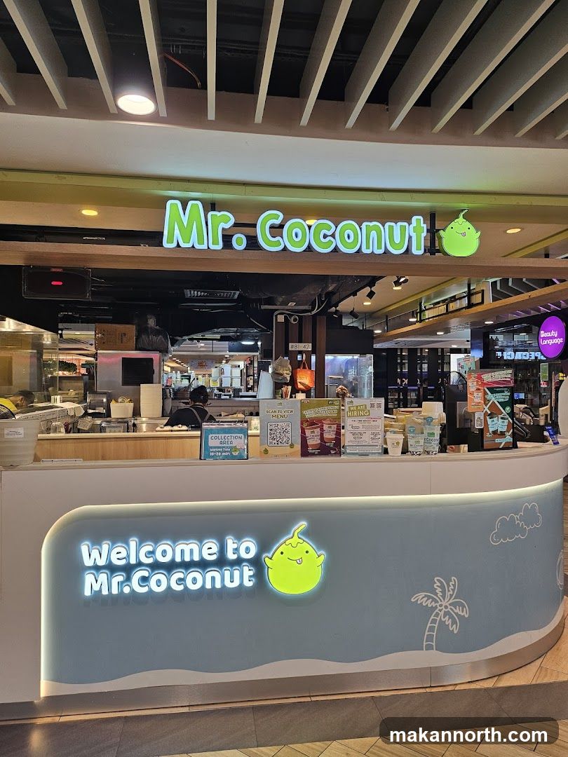 A brightly lit storefront for Mr. Coconut, a business that appears to sell coconut-based drinks and desserts. The prominent signage above the counter reads "Mr. Coconut" in green lettering, accompanied by the brand's cartoon coconut mascot. The counter itself has a blue and white design with "Welcome to Mr. Coconut" illuminated in white text and the mascot. Behind the counter, staff members are visible preparing orders, and there are various displays and signs, including a QR code for viewing the menu and a "We are Hiring" poster.
