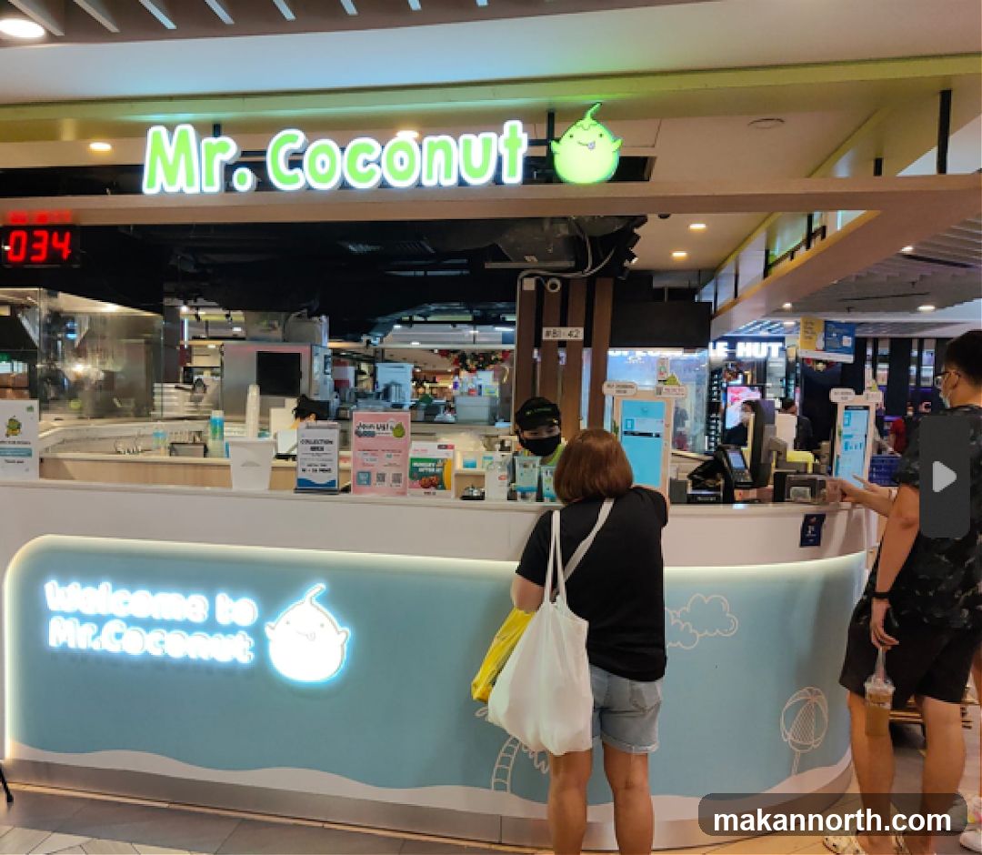 A brightly lit storefront for Mr. Coconut, a business that appears to sell coconut-based drinks and desserts. The signage above the counter clearly displays the brand name "Mr. Coconut" with a cartoon coconut logo. The counter itself is decorated with a light blue wave pattern and the words "welcome to Mr. Coconut" in illuminated lettering. Several customers are interacting with staff at the counter, and digital ordering screens are visible. The overall impression is a modern and inviting dessert shop.