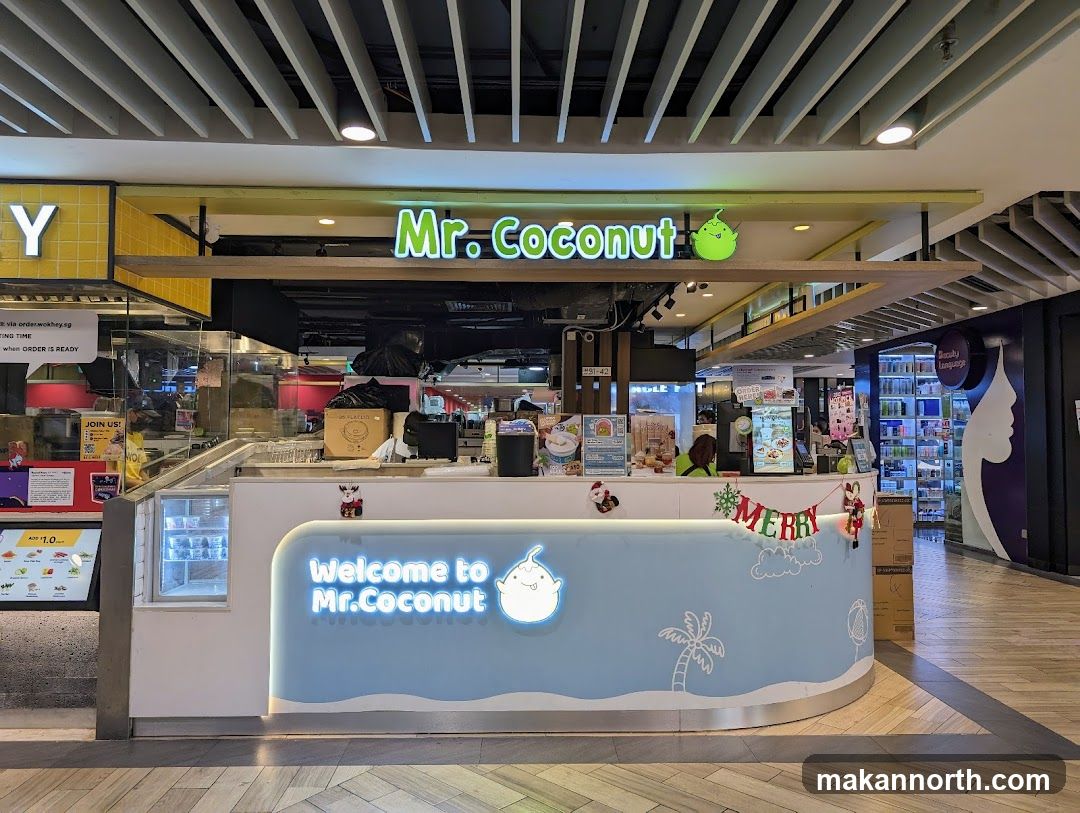 The exterior of a Mr. Coconut store, a dessert shop, located in a mall. The store has a light blue counter with "Welcome to Mr.Coconut" written in neon blue. The store's name "Mr. Coconut" is also visible in neon green above the counter. There are Christmas decorations on the counter.