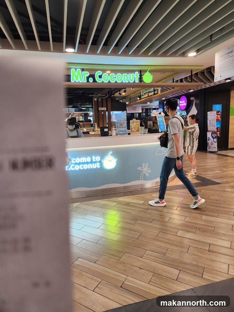 A brightly lit storefront for "Mr. Coconut" with neon green signage. A counter is visible with staff members serving customers. The counter has a light blue base with a cartoon coconut character and palm tree graphic. Two customers are walking past the storefront. The floor is made of light-colored wood planks.