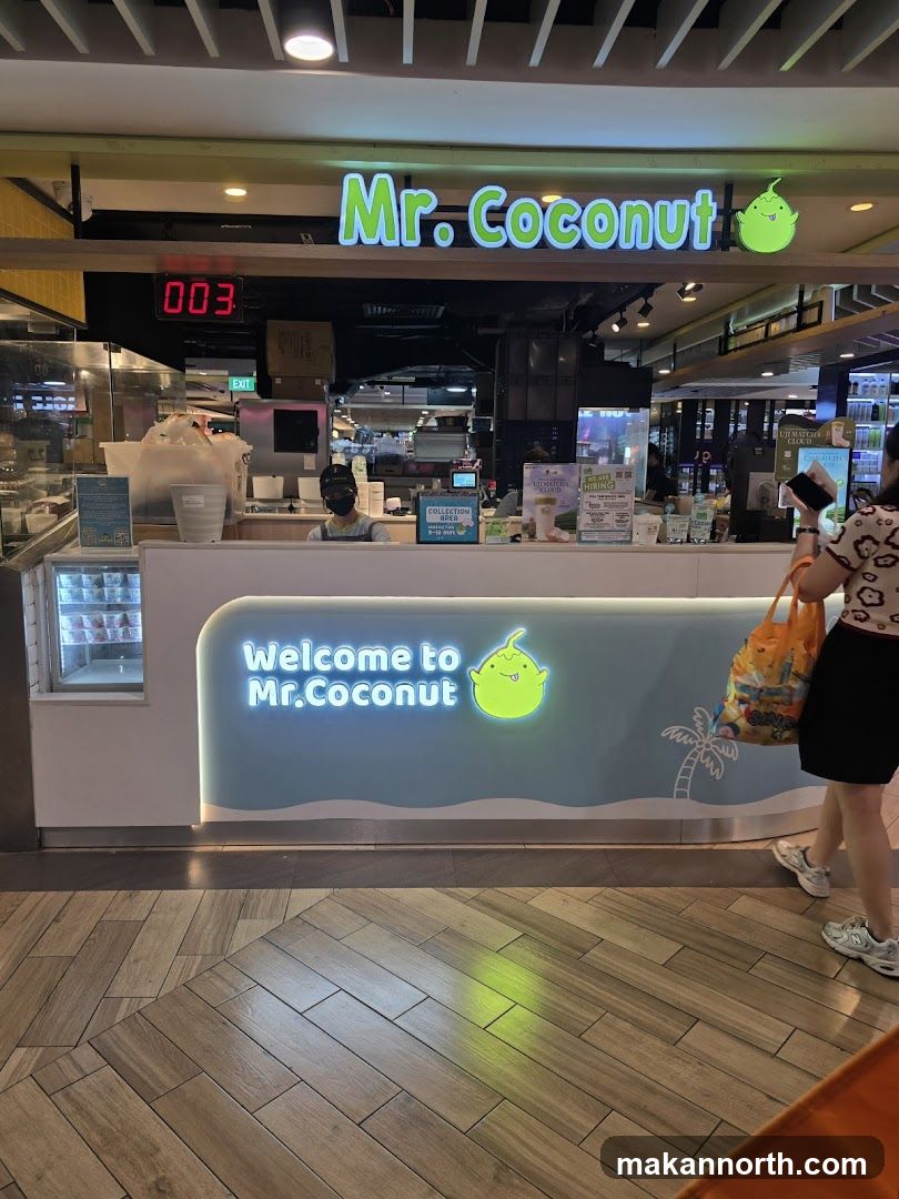 A brightly lit storefront for "Mr. Coconut" with a prominent neon sign. The counter area is visible, with a staff member behind it. A digital display shows "003". The counter has a blue wave design with the text "Welcome to Mr. Coconut" and a cartoon coconut logo. A customer is walking past the storefront carrying a colorful shopping bag.
