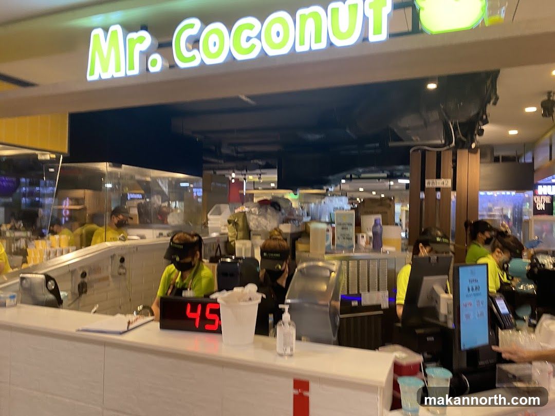 A view of the Mr. Coconut store counter with employees preparing orders. The prominent "Mr. Coconut" signage is visible above the counter. Several employees wearing green shirts and black caps are working behind the counter. A digital display shows "45". There are also cups, a hand sanitizer dispenser, and a payment terminal on the counter. In the background, more store operations and customers are visible.