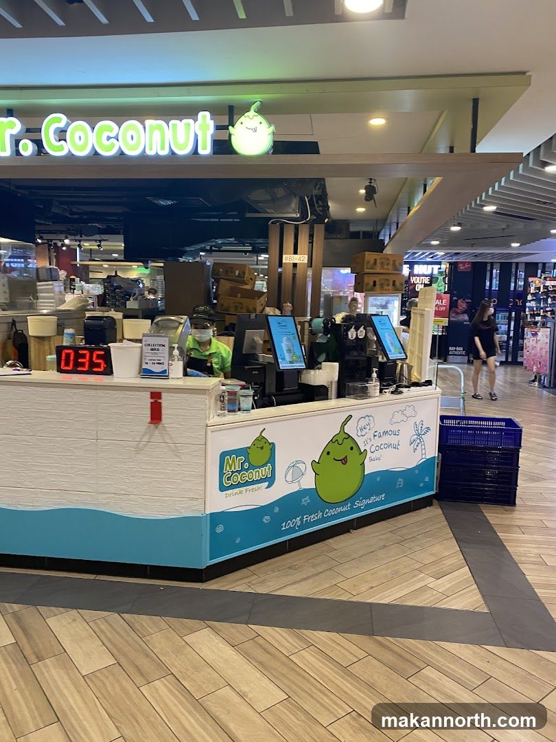A Mr. Coconut stall with a prominent sign and cartoon coconut branding. The stall has a white counter with a blue wave design and a digital display showing '035'. Staff members are visible behind the counter, and there are screens displaying information. In the background, there are shelves and other retail displays.