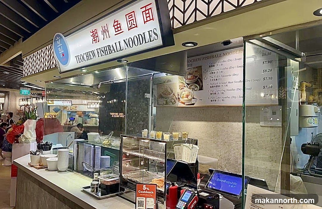 A food stall with a sign that reads "Teochew Fishball Noodles" in English and Chinese. A menu is displayed on the glass counter, listing various noodle dishes and their prices. The stall appears to be in a food court setting, with other stalls visible in the background.