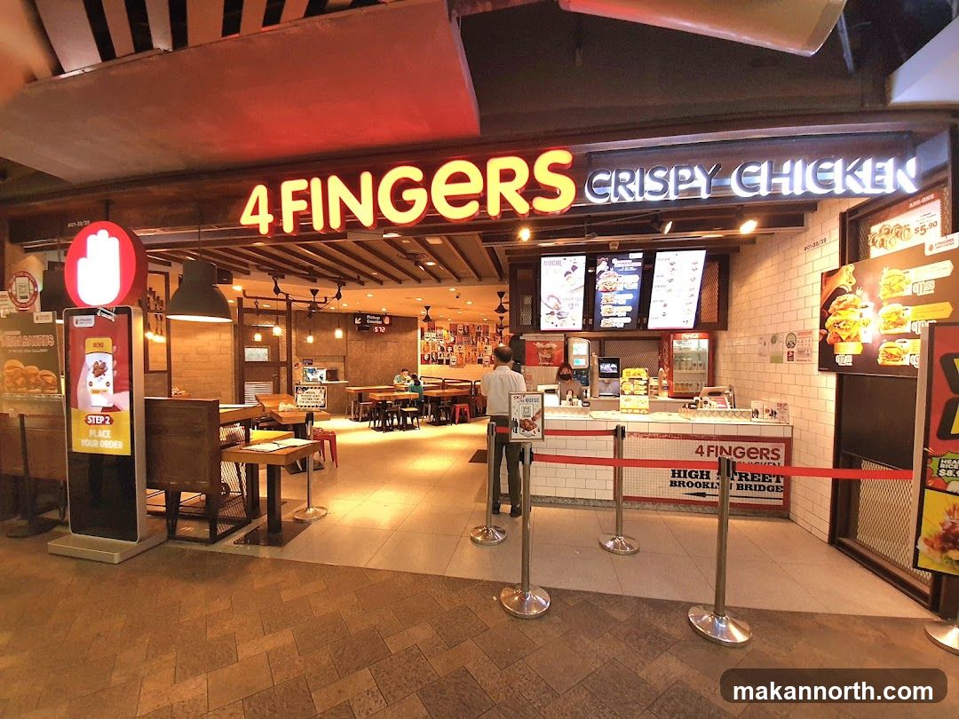 The exterior of a 4 Fingers Crispy Chicken restaurant. The storefront features a large neon sign that reads "4 FINGERS CRISPY CHICKEN". There is a digital ordering kiosk to the left of the entrance and a counter with menu boards above it. Red velvet ropes guide customers into the restaurant.