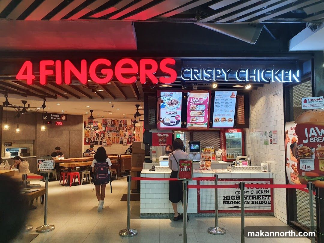 The exterior of a 4 Fingers Crispy Chicken restaurant. The prominent red neon sign reads "4 FINGERS" and a white neon sign reads "CRISPY CHICKEN". There are menu boards displayed, showing various chicken and burger options. Inside, customers are seated at tables, and staff are visible behind the counter. A pickup counter with a digital display showing "389" is also visible.