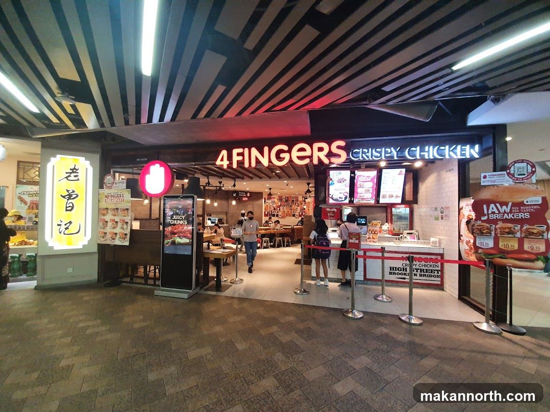 The exterior of a 4 Fingers Crispy Chicken restaurant. The storefront features the restaurant's name in large neon letters above the entrance. There are menu boards and promotional displays visible, including a large advertisement for "Jaw Breakers" burgers. A digital display shows "Juicy Chunks." Several people are seen inside and queuing outside.