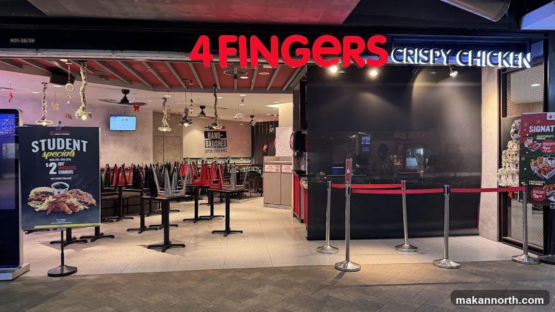 The exterior of a 4 Fingers Crispy Chicken restaurant. Red neon signs spell out "4 FINGERS" above the entrance, with "CRISPY CHICKEN" in white neon to the right. Inside, tables and chairs are stacked, suggesting the restaurant is closed or preparing to close. A promotional poster for "STUDENT specials" is visible to the left, offering $2 off combos. Red velvet ropes guide customers towards the entrance.