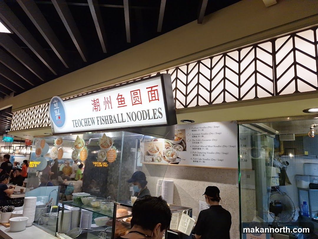 A brightly lit stall with a sign that reads "Teochew Fishball Noodles" in English and Chinese. Below the sign, a menu lists various noodle dishes with prices. The stall appears to be in a food court setting with other stalls and patrons visible in the background.