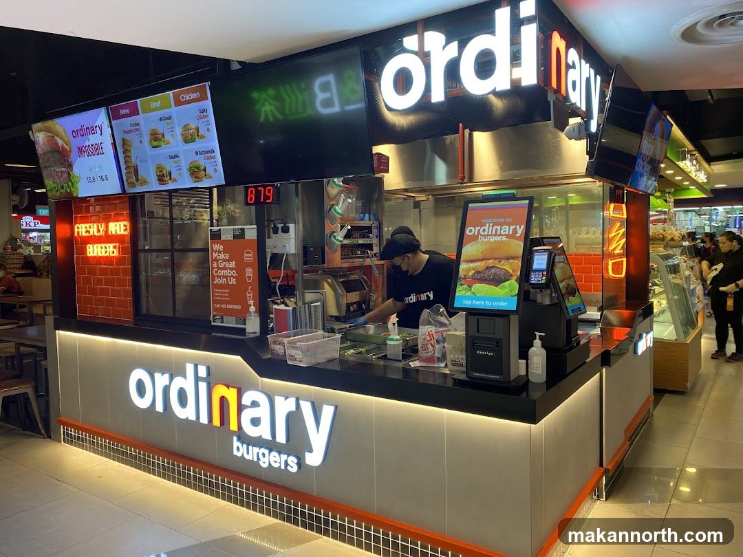 A brightly lit burger stall with the "ordinary burgers" logo prominently displayed on the front counter and above the stall. Digital menus show various burger options. A staff member is visible preparing food behind the counter.