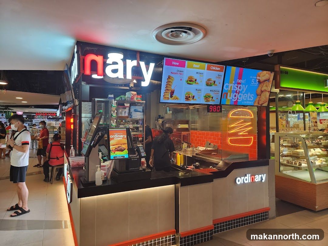 A brightly lit fast-food counter with a prominent "ordinary" logo and neon burger sign. Digital menu boards display various burger and chicken nugget options. Customers are visible in the foreground and background.