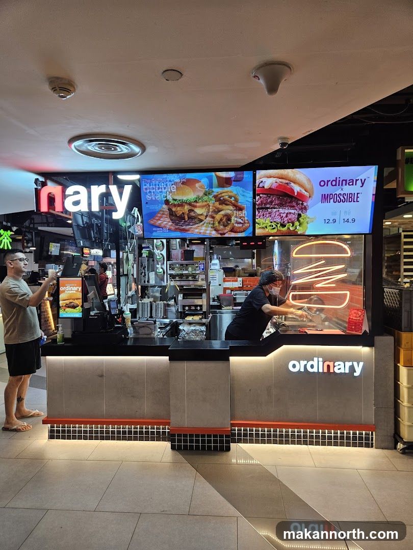 A burger stand with the name "ordinary" displayed prominently on a neon sign and a backlit panel. Two digital menu boards are visible, one showing a classic double stock burger and onion rings, and the other featuring an Impossible burger. Two employees are working behind the counter.