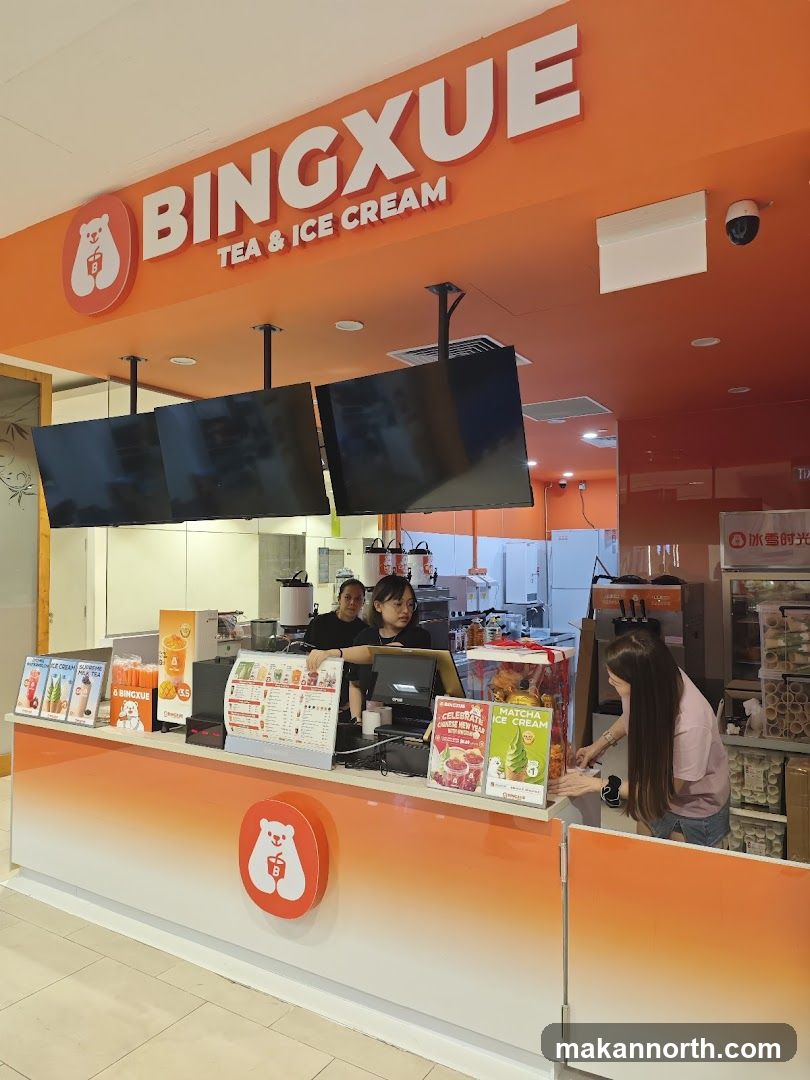 The storefront of a Bingxue Tea & Ice Cream shop. The prominent orange signage above the counter reads "BINGXUE TEA & ICE CREAM" with a polar bear logo. Inside, staff members are visible behind the counter, serving customers. Various promotional materials and menus are displayed on the counter.