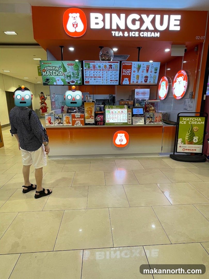 The exterior of a Bingxue Tea & Ice Cream shop with a prominent orange facade and illuminated signage. Digital menu boards display various drinks and ice cream options, including "Matcha" and "All-Time Top Picks." A person with a robot emoji face is standing in front of the counter. A promotional sign for "Matcha Ice Cream" is visible to the right.