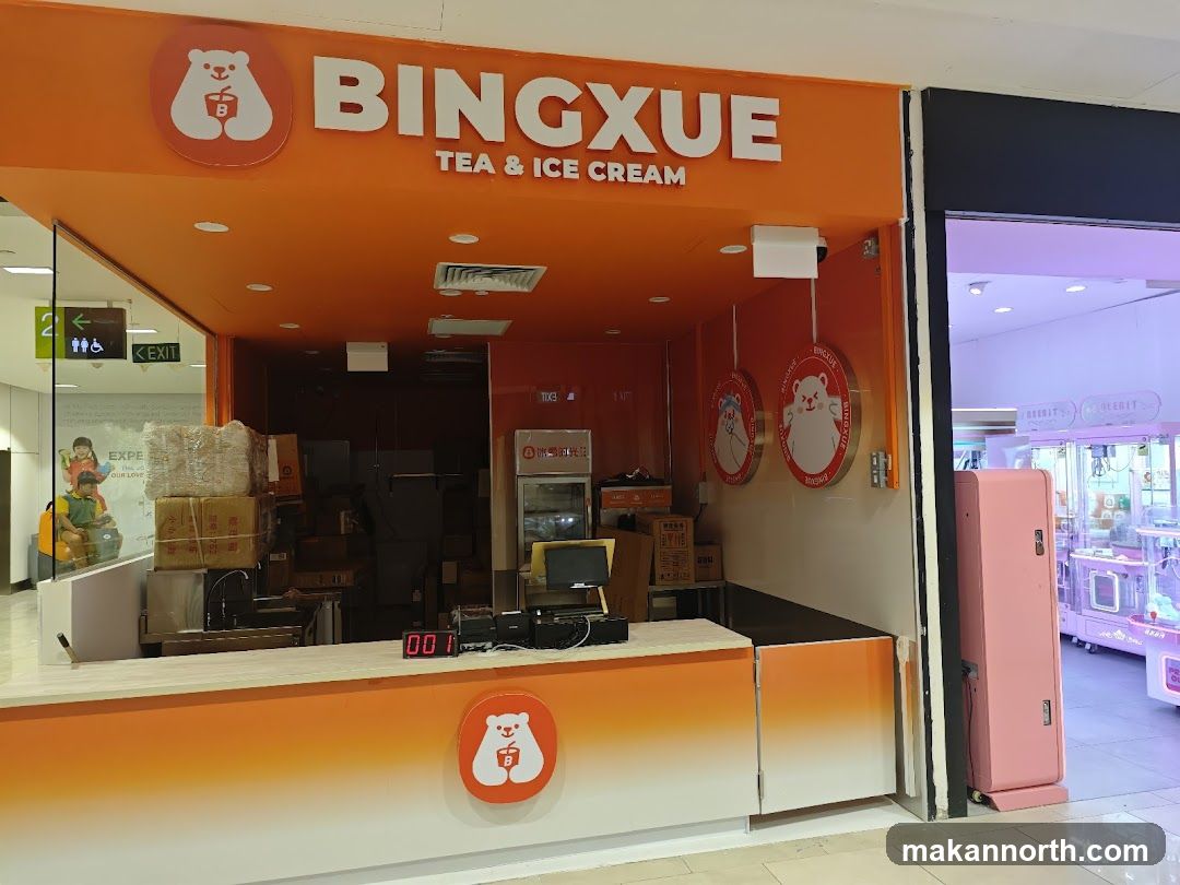The exterior of a Bingxue Tea & Ice Cream shop. The shop has an orange facade with the business name and logo prominently displayed. Inside, there are stacks of boxes and a counter with a digital display. To the right, a pink vending machine and arcade games are visible.