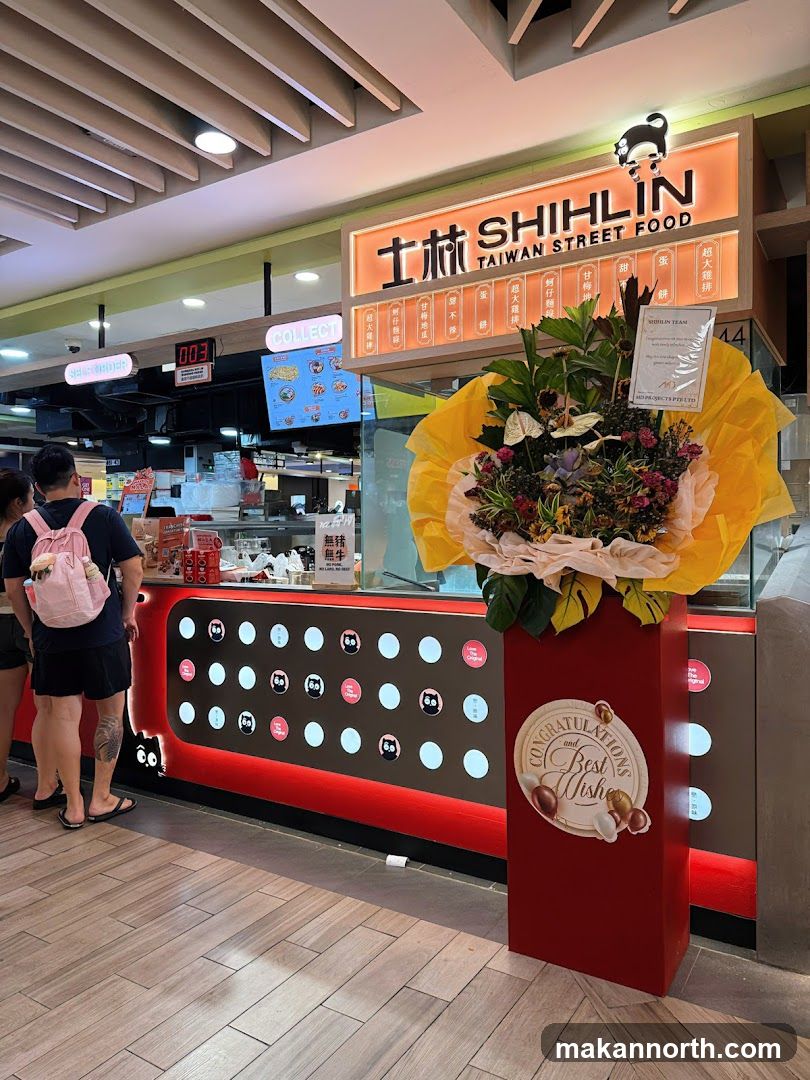 A Shihlin Taiwan Street Food storefront with a large illuminated sign featuring the brand name and logo. Below the sign, a counter displays various food items and a menu board. A bouquet of flowers is placed on a red pedestal in front of the counter.