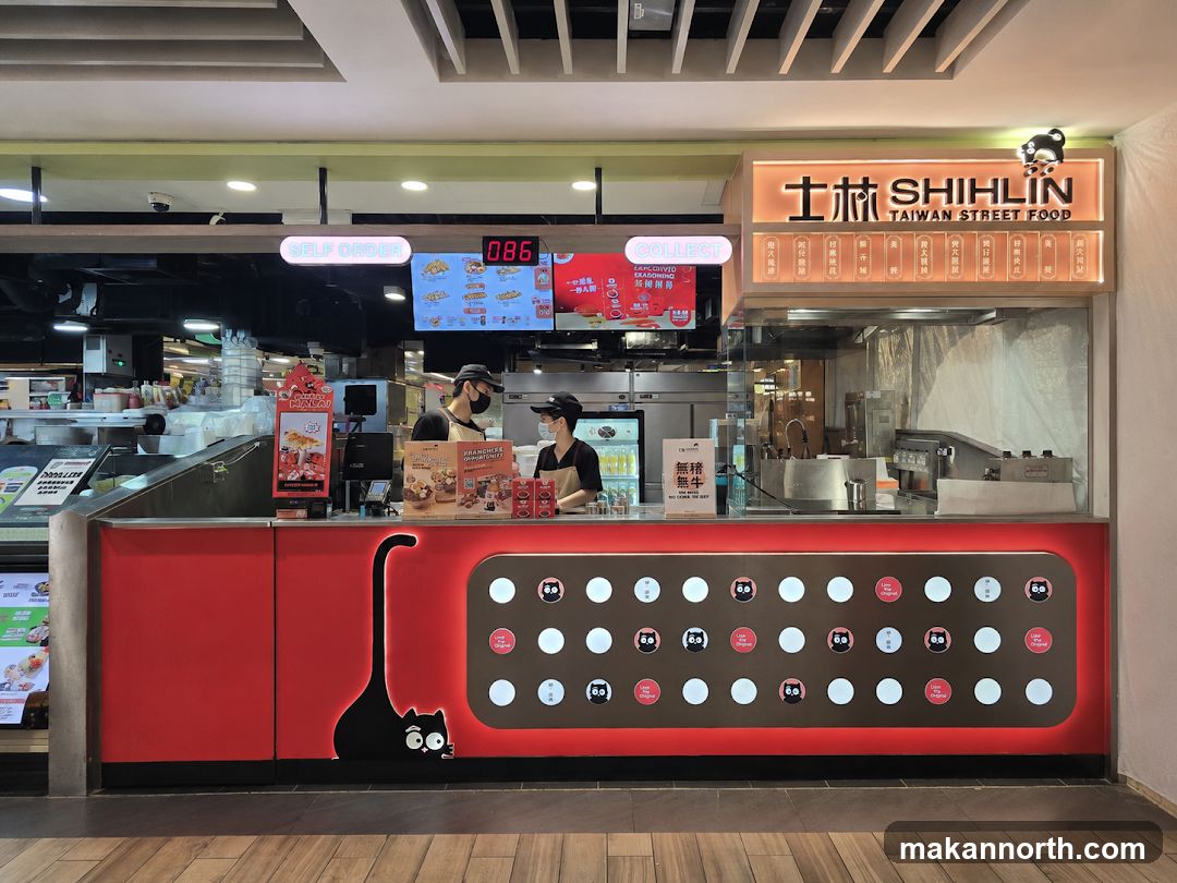 A Shihlin Taiwan Street Food stall with a prominent sign featuring the business name and logo. The stall has a red counter with a black cat graphic. Digital menu boards are visible above the counter, displaying food items and prices. Two employees in black uniforms and caps are standing behind the counter.