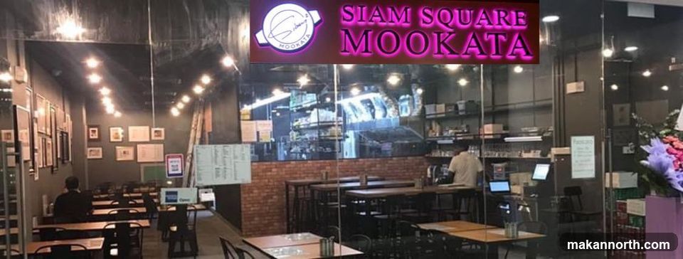 The exterior of a restaurant with a prominent neon sign that reads "SIAM SQUARE MOOKATA". The entrance features large glass windows and doors, offering a view into the dining area with tables and chairs.