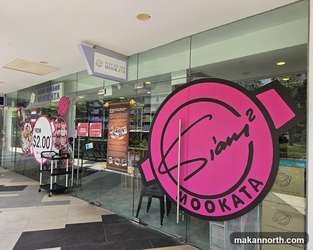 The exterior of a restaurant with large glass windows and doors. A prominent pink circular logo with the text "Siam² Mookata" is displayed on the right door. A directional sign above the entrance reads "Siam Square Mookata". Various promotional posters and images are visible through the glass, including one advertising a deal starting from $2.00.