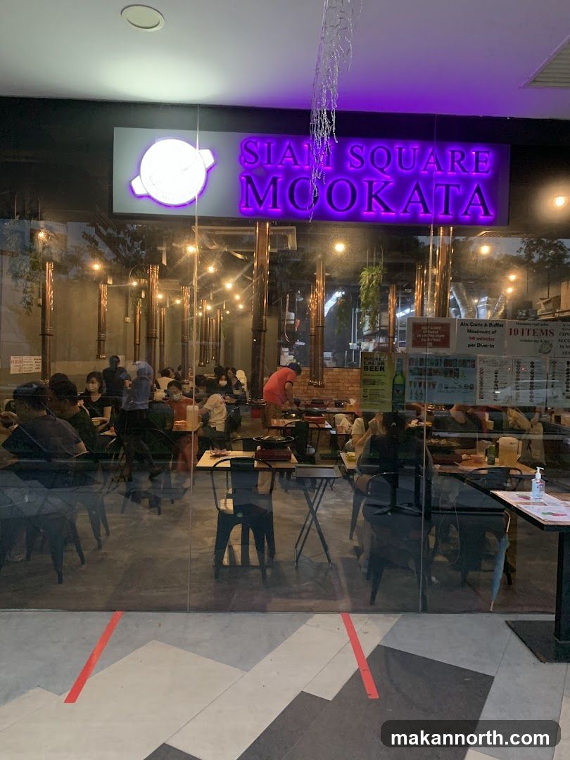 The exterior of a restaurant with a large illuminated sign that reads "SIAM SQUARE MOOKATA". Through the glass windows, diners can be seen seated at tables with cooking equipment. The interior is lit with warm lighting and hanging decorative lights.