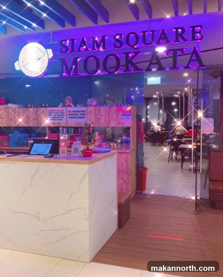 The exterior of a restaurant with a prominent sign reading "SIAM SQUARE MOOKATA" in large, illuminated letters. The entrance is visible, leading into a dining area with tables and chairs. A counter is positioned near the entrance, with a computer monitor and various items on display. The overall ambiance is enhanced by sparkling light effects.