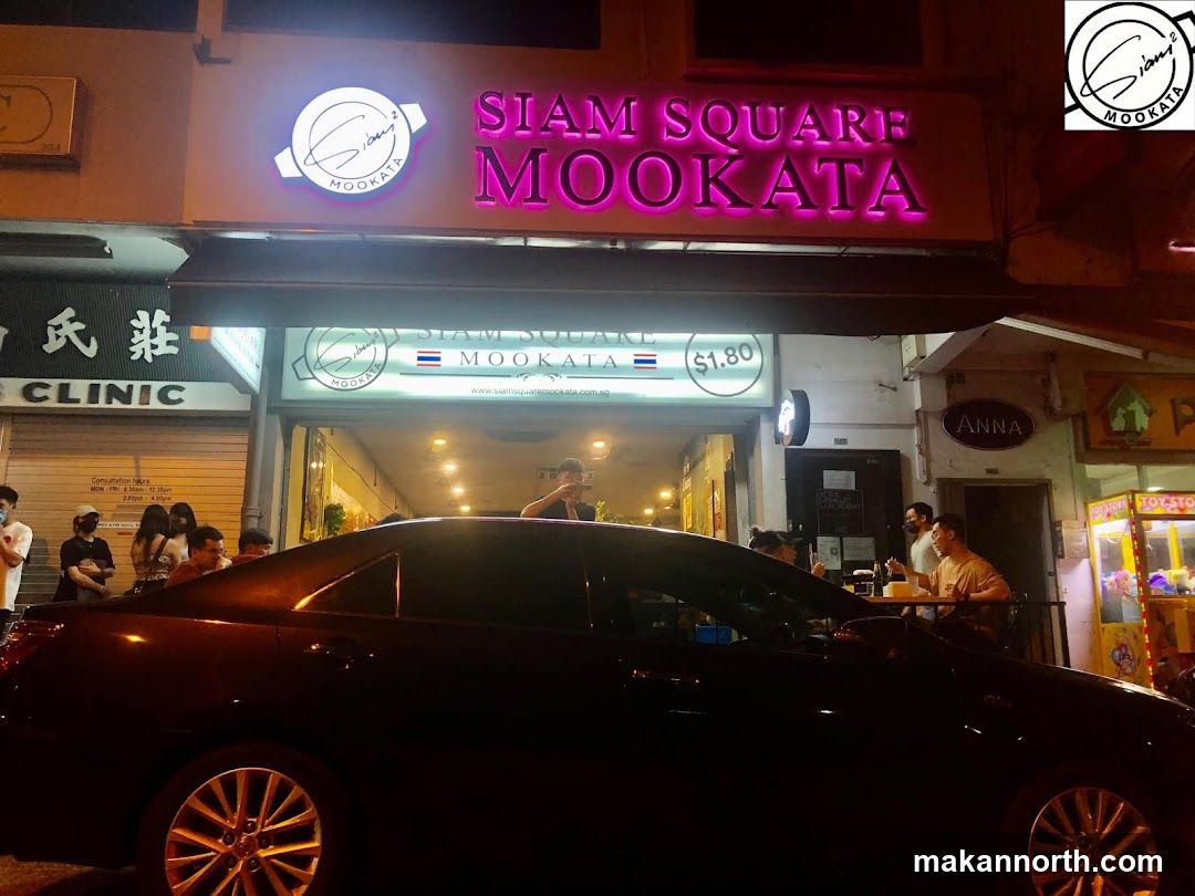 A neon sign with the words "SIAM SQUARE MOOKATA" in pink letters is visible above the entrance of a restaurant. Below this, a smaller sign reads "SIAM SQUARE MOOKATA" with a Thai flag and "$1.80". People are seen inside the restaurant, and a car is parked in front.
