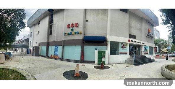 A panoramic view of the exterior of the 600 Toa Payoh building. The building has a light-colored facade. On the left side, there is a sign with colorful letters that reads "600 TOAPAYOH". To the right, there is an entrance to the Bank of China with stairs leading up to it. A McDonald's sign is visible on the upper right side of the building. The area in front of the building appears to be a public walkway with some trees and planters.