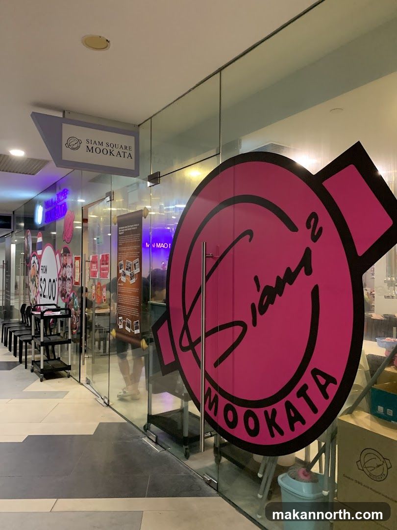 The exterior of a restaurant with large glass windows and doors. A prominent pink circular logo with the text "Siam Square Mookata" is visible on the glass. A smaller sign above the entrance also reads "Siam Square Mookata". Inside, chairs and tables are visible, along with some promotional materials and food items.