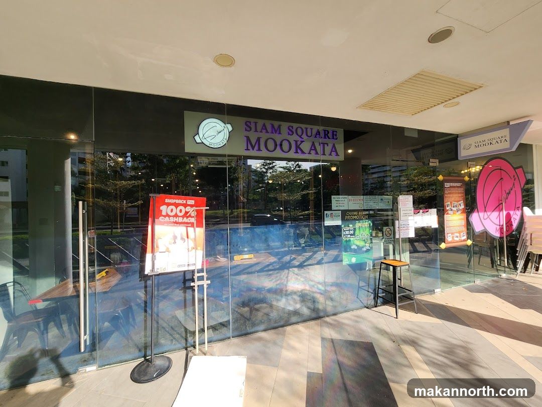 The exterior of a restaurant with large glass windows and doors. A prominent sign above the entrance reads "SIAM SQUARE MOOKATA". Various promotional posters and signs are displayed on the glass, including one advertising "100% CASHBACK" and another for a "CHANG BEER PROMOTION". Inside, tables and chairs are visible.