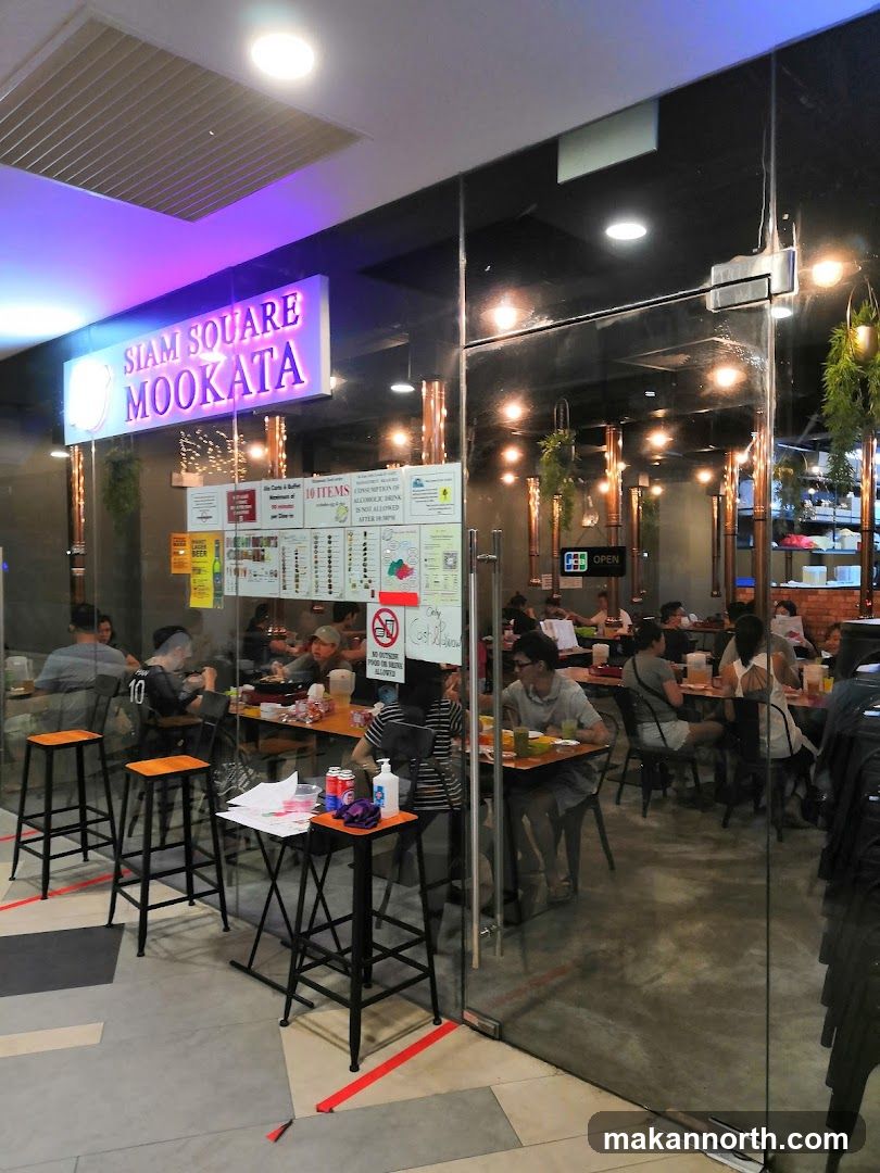 The exterior of a restaurant called Siam Square Mookata. The restaurant has large glass doors and windows, allowing a view of the interior where diners are seated at tables. A prominent sign above the entrance reads "SIAM SQUARE MOOKATA" in illuminated purple and white lettering. Inside, the restaurant appears to be busy with customers.