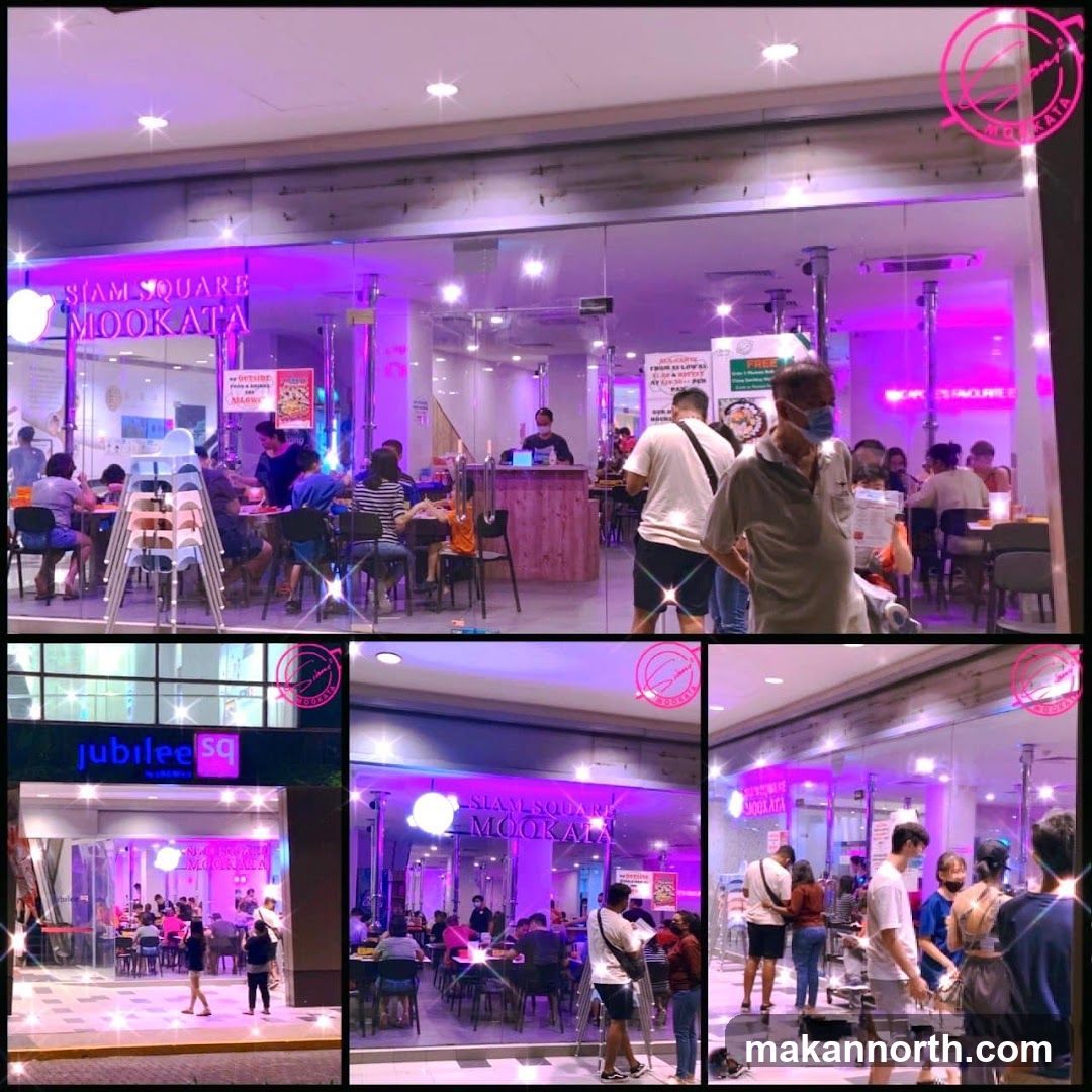 The exterior of a restaurant with large glass windows and doors. The restaurant's name, "Siam Square Mookata," is visible on the signage above the entrance. Inside, customers are seated at tables, and staff are visible behind a counter. The interior is illuminated with purple lighting.