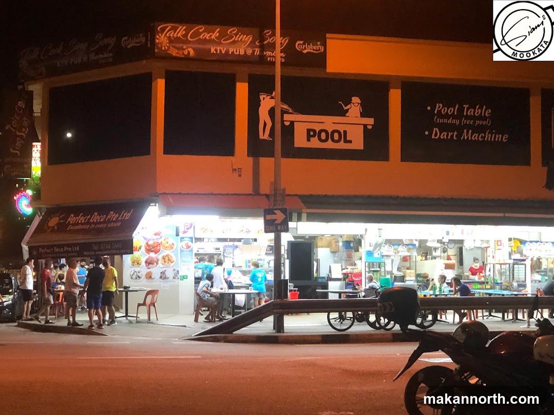 A street-level view of a KTV pub and restaurant establishment at night. The upper facade features signage for "Talk Cook Sing Song" and "Carlsberg". Below, the establishment has a large window displaying food items and seating areas. A separate awning displays "Perfect Deco Pte Ltd" with contact information. People are seen dining and socializing outside and inside the establishment.