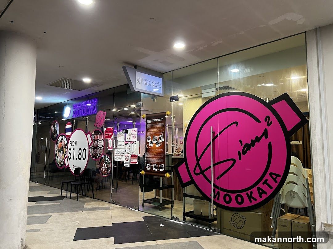 The exterior of a restaurant with large glass windows and doors. A prominent pink circular logo with the text "Siam Square Mookata" is visible on the right side of the entrance. Above the entrance, a sign reads "SIAM SQUARE MOOKATA". Inside, tables and chairs are visible, along with some menu boards and promotional materials.