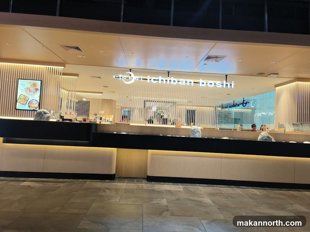 The exterior of a modern Japanese restaurant with a clean and minimalist design. The restaurant's name, "Ichiban Boshi," is displayed in illuminated white lettering above the entrance. The facade features vertical wooden slats and a dark counter area. A digital menu board with images of food is visible to the left.