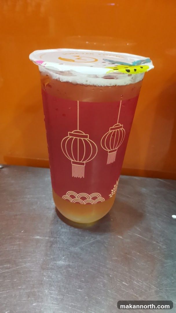 A close-up shot of a beverage in a clear plastic cup with a red sleeve. The sleeve is decorated with white outlines of lanterns and a wave pattern at the bottom. The drink appears to have two layers: a lighter colored liquid at the bottom and a darker, reddish liquid on top, topped with foam. The cup is sealed with a white lid, and a yellow tab with a smiley face design is attached to the lid. The background is a solid orange color, and the cup is resting on a metallic surface.