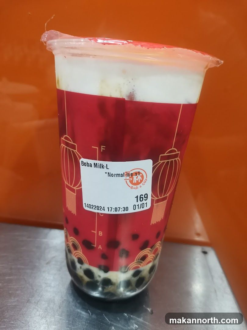 A plastic cup filled with boba milk tea. The cup has a red sleeve with a pattern of Chinese lanterns and decorative waves at the bottom. The drink contains milk, tea, and tapioca pearls. A white label on the cup reads "Boba Milk-L", "*Normal Ice x1", and has a price of "169". Below that, it shows a date and time "14022024 17:07:30" and another date "01/01". There are also markings "A" and "B" on the cup.